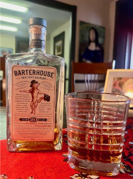 Bottle of Barter House bourbon beside a glass of whiskey on a red table